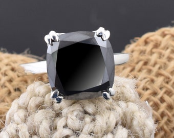 May include: A silver ring featuring a large, square-cut black gemstone. The gemstone is set with four prongs. The ring is displayed on a textured, beige fabric background. The ring's band is a simple, polished silver.