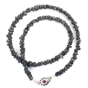 Pode incluir: Um colar de diamantes negros com um fecho prateado que apresenta uma pequena pedra vermelha. O colar é feito de diamantes negros brutos e não cortados.
