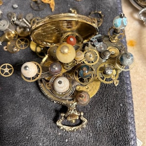 May include: A steampunk-style assemblage of watch parts and decorative elements. The composition features a gold-colored pocket watch case, numerous gears, and various spherical embellishments in shades of white, blue, and brown. The arrangement is set against a dark gray backdrop.