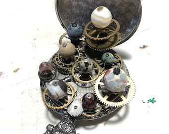 Può includere: Un orologio da tasca in argento con un coperchio incernierato aperto, che rivela un complesso meccanismo di ingranaggi in ottone e sfere di agata. L'orologio ha un'impugnatura decorativa.