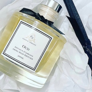 Puede incluir: Una botella de vidrio transparente con una cinta negra y una tapa plateada. La botella contiene un líquido amarillo claro y tiene una etiqueta que dice "Chateau Ambiance OUD Diffuseur d'Ambiance Aroma Reed Diffuser 200ml."