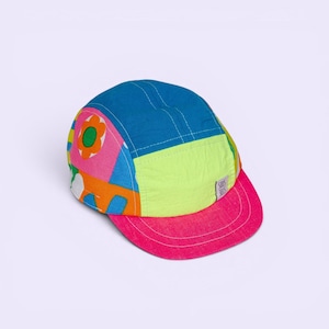 May include: A colorful five-panel cap with a vibrant design. The cap features a blue top, a neon green side panel, and a pink brim. The design includes a floral pattern with pink, orange, and white accents. The cap has a small white tag on the side.