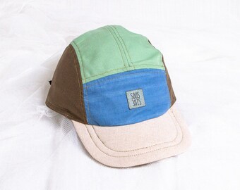 May include: A multi-colored five-panel cap with a flat brim. The cap features panels in green, brown, and blue, with a beige brim. A small rectangular label with the text "SOUS 3015" is sewn on the blue panel. The cap is likely made of fabric.