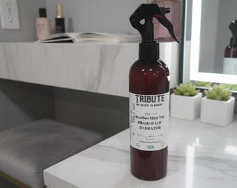 Op de afbeelding: Een bruine spuitfles met een zwarte spuitkop en een wit etiket met de tekst "TRIBUTE" en "Rooibos Mint Tea Braid & Loc Hydrator". De fles staat op een wit aanrechtblad met een marmerpatroon. De achtergrond omvat een kaptafel en een spiegel.