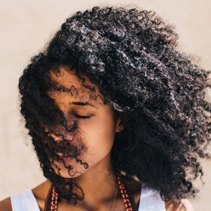 May include: A woman with long, curly, black hair. Her hair is covering most of her face. She is wearing a white tank top and a beaded necklace.