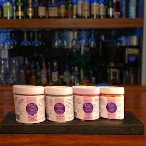 May include: Four jars of bath products with white lids and purple labels sit on a dark wooden tray. The jars contain colorful bath products in shades of orange, pink, green, and brown. The background includes a bar with bottles and glasses.