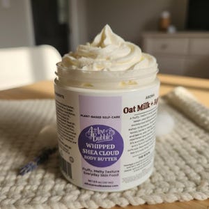May include: A jar of whipped shea cloud body butter with a fluffy, melty texture. The jar has a white label with purple accents and the text "The Love of Bubbles". The product is plant-based and has an Oat Milk + Agave aroma.