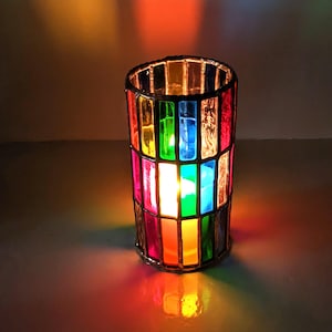 May include: A colorful stained glass candle holder with a lit candle inside. The holder is made of glass and has a rainbow of colors, including red, orange, yellow, green, blue, and purple.