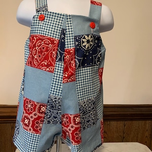 Könnte beinhalten: Ein ärmelloser Strampler mit einem Patchwork-Design aus roten, weißen und blauen Bandana-Mustern, kombiniert mit hellblau-weißem Vichy-Karo. Das Kleidungsstück verfügt über zwei leuchtend rote Knöpfe an der Oberseite der vorderen Träger.