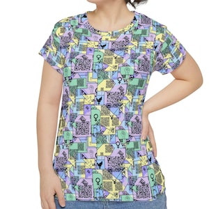 Puede incluir: Una camiseta de manga corta con un colorido estampado geométrico. El diseño presenta una mezcla de colores pastel, incluyendo amarillo, verde y morado, con códigos QR y otros símbolos. La camiseta se lleva con vaqueros azules.