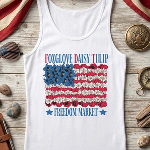 White tank top with American flag design featuring "Foxglove Daisy Tulip" and "Freedom Market".