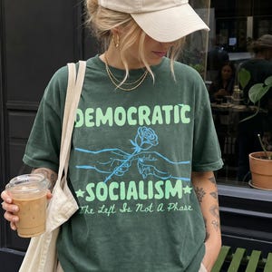 May include: A dark green short-sleeve t-shirt featuring a light green graphic with the text DEMOCRATIC SOCIALISM and The Left Is Not A Phase, along with a blue illustration of two hands holding a rose, paired with a beige baseball cap.
