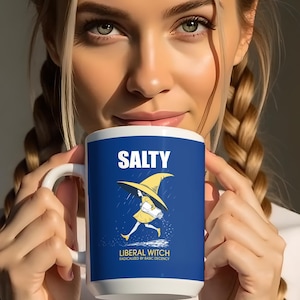 May include: White ceramic mug with a blue background featuring the word "SALTY" at the top. Below is a cartoon of a witch running, and the text "LIBERAL WITCH" is at the bottom. The mug is held by a person.