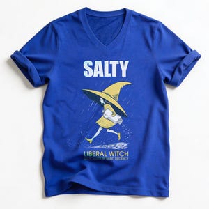 May include: Royal blue v-neck t-shirt with rolled-up sleeves. The shirt features a graphic of a witch in a yellow dress and hat, running in the rain, with the word "SALTY" above and "LIBERAL WITCH" below.