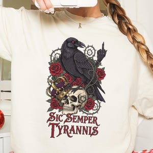 May include: Cream-colored t-shirt featuring a graphic design with a black raven, red roses, a skull wearing a crown, and the text "Sic Semper Tyrannis". The raven is perched above the skull, with a middle finger raised. The design is surrounded by thorns.