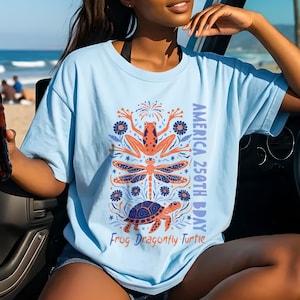 Puede incluir: Camiseta azul claro con un diseño de rana, libélula y tortuga. El texto "AMERICA 250TH BDAY" y "Frog Dragonfly Turtle" están impresos. Una persona sostiene una botella de líquido rojo.