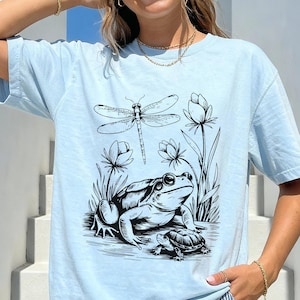 May include: Light blue t-shirt featuring a black and white illustration of a frog, dragonfly, turtle, and flowers. The word "Chambray" is printed in the bottom right corner. The shirt has short sleeves and a relaxed fit.