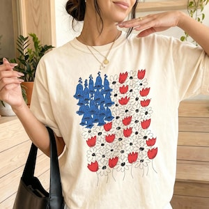 May include: A cream-colored short-sleeve t-shirt featuring a graphic print of blue bell-shaped flowers and red and white daisies arranged in a rectangular pattern. The shirt is paired with a white pleated skirt and a black shoulder bag.