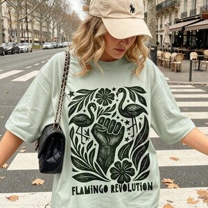 May include: A light green t-shirt featuring a black graphic print of two flamingos, a raised fist, and floral elements, with the text "FLAMINGO REVOLUTION" printed at the bottom. The shirt is paired with a beige baseball cap and a black shoulder bag.