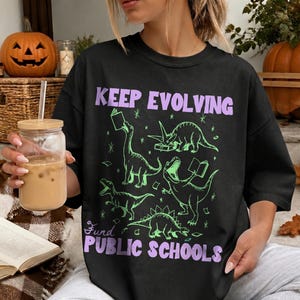 May include: A black t-shirt featuring a graphic print of five green dinosaurs reading books, with the purple text "KEEP EVOLVING" at the top and "FUND PUBLIC SCHOOLS" at the bottom, surrounded by small star accents.
