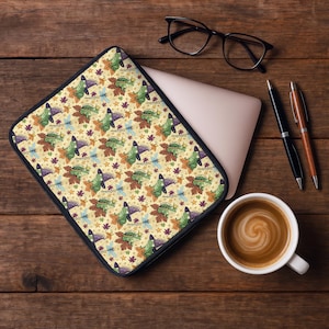 May include: A laptop sleeve with a whimsical pattern featuring green cartoon dinosaurs wearing witch hats, surrounded by autumn leaves and dragonflies on a cream background. The sleeve is on a wooden desk with a laptop, coffee, and pens.
