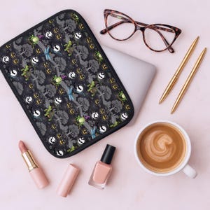 May include: A black laptop sleeve with a Halloween-themed pattern featuring trees, pumpkins, and dragonflies. The image also includes a laptop, glasses, pens, lipstick, nail polish, and a cup of coffee, all arranged on a pink surface.