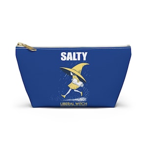 May include: A blue zippered pouch with a gold zipper pull. The pouch features the word "SALTY" in white, above an illustration of a witch in a yellow hat and dress, running in the rain. The text "LIBERAL WITCH" is below the illustration.