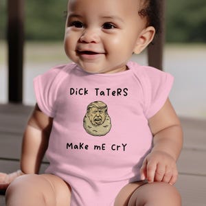 May include: A baby wearing a light pink onesie with the text "Dick TaTeRs" above an illustration of a potato with a face. Below the illustration is the text "Make me CrY". The baby is sitting on a wooden surface.