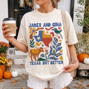 May include: A cream-colored short-sleeve t-shirt featuring a colorful graphic print with the text "JAMES AND GINA" and "TEXAS. BUT BETTER." The design includes a map of Texas, a longhorn, a bird, and floral elements in blue, orange, and green.