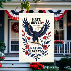 May include: A cream garden flag with a bald eagle design, red flowers, and blue leaves. The flag has the text "Hate Never Made Any Nation Great". The flag is displayed on a black metal stand, suitable for a garden.