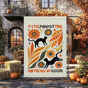 May include: A rectangular garden flag with a cream background featuring two black cats, orange and olive green flowers, and abstract leaf shapes. The text reads IT'S THE PURRFECT TIME FOR THE FALL OF FASCISM in orange and black lettering.
