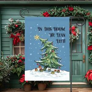 May include: A blue Christmas garden flag with the text "IN THIS HOUSE WE LEAN LEFT!" features a decorated Christmas tree with ornaments and gifts on a snowy ground. Snowflakes are falling in the background.