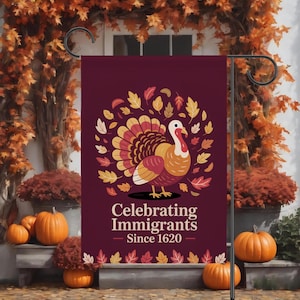 May include: A burgundy garden flag with a turkey illustration and autumn leaves. The text reads "Celebrating Immigrants Since 1620." Pumpkins and fall foliage are in the background. The flag is displayed outdoors.