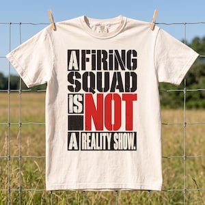 May include: A cream-colored short-sleeve t-shirt featuring bold, distressed black and red text that reads A FIRING SQUAD IS NOT A REALITY SHOW. The graphic is centered on the chest, utilizing a blocky, stencil-style font with a worn texture.