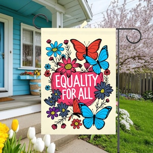 May include: A garden flag displaying the words "EQUALITY FOR ALL" in white on a pink heart-shaped background. The flag features illustrations of red, blue, and orange butterflies and colourful flowers. The flag is displayed on a black metal stand.