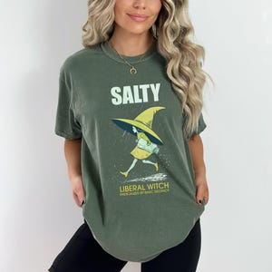 Pu&ograve; includere: T-shirt verde oliva con la scritta "SALTY" sopra una grafica di una strega con cappello e vestito gialli che corre sotto la pioggia. Sotto c'&egrave; la scritta "LIBERAL WITCH RADICALIZED BY BASIC DECENCY".