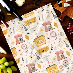 May include: A cream-colored kitchen towel with a repeating pattern of pink stand mixers, yellow slow cookers, clocks, and floral designs. The text "FUCK TRUMP" is also visible. The towel is surrounded by wine bottles, cheese, and grapes.