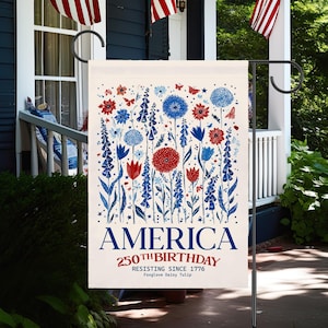 May include: A garden flag with a floral design in red, white, and blue. The flag reads "AMERICA 250th BIRTHDAY RESISTING SINCE 1776" with the words "Foxglove Daisy Tulip" below. The flag is displayed on a black metal stand.