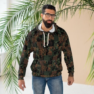 May include: A long-sleeve hooded sweatshirt featuring a dark camouflage pattern composed of various square patches. The design includes repeating symbols such as raised fists, gender icons, and QR codes in shades of brown, green, and black.