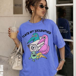 May include: A periwinkle blue crewneck t-shirt featuring a graphic print of a security camera surrounded by a teal bird, a pink bird, a yellow bird, and a small green creature, with the text FLOCK OFF BIGBROTHER printed in black above the graphic.