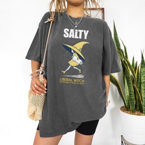 Pu&ograve; includere: T-shirt grigio scuro con la scritta "SALTY" sopra una grafica di una strega con abito e cappello gialli. Il testo "LIBERAL WITCH Radicalized by Basic Decency" &egrave; sotto. La maglietta &egrave; oversize.