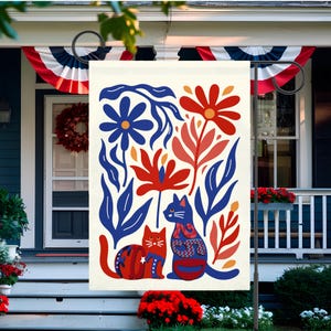 May include: A cream-colored garden flag featuring a whimsical design with two cats and floral elements in red, blue, and orange. The cats are adorned with decorative patterns. The flag is displayed on a black metal stand, perfect for outdoor decor.