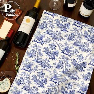 Puede incluir: Un paño de cocina blanco y azul estampado con cerdos voladores, árboles y edificios. El texto "PIGGY RESISTANCE" y "FUCK TRUMP" está impreso en el paño. Botellas de vino, queso y una tabla de cortar en el fondo.
