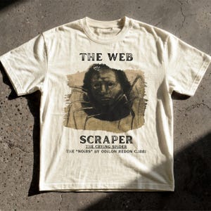 May include: An off-white short-sleeve t-shirt featuring a graphic print of a spider with a human face. The text reads THE WEB, SCRAPER, THE CRYING SPIDER, and THE NOIRS BY ODILON REDON C.1881. The design is centred on the chest of the garment.