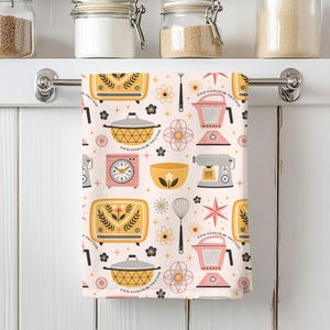 May include: A kitchen towel with a repeating pattern of retro kitchen appliances and floral designs in yellow, pink, and gray on a light pink background. The towel includes images of a toaster, mixer, and clock.