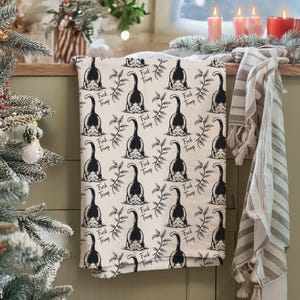 May include: A cream-colored kitchen towel with a repeating pattern of a black cat's rear end and the words "Fuck Trump" in black. The towel also features a sprig of leaves.