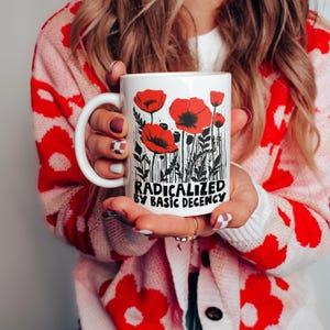 May include: White ceramic mug featuring a floral design of red poppies with black stems and leaves. The mug has the text "RADICALIZED BY BASIC DECENCY" in black. The mug is held by a person wearing a red and white floral cardigan.
