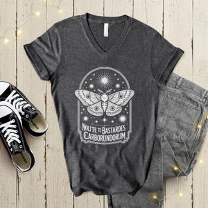 May include: Dark gray V-neck t-shirt featuring a white moth design within a decorative frame, with the text "Nolite Te Bastardes Carborundorum". The shirt is paired with black and white sneakers and denim jeans.