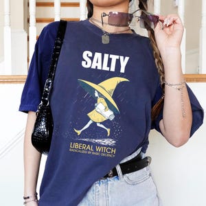 May include: A navy blue short-sleeve t-shirt featuring a graphic of a person in a yellow witch hat walking in the rain. The text SALTY is printed in white above the graphic, and LIBERAL WITCH RADICALIZED BY BASIC DECENCY is printed in yellow below.