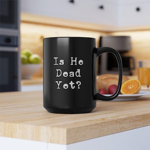 May include: Black ceramic coffee mug with white text that reads "Is He Dead Yet?"